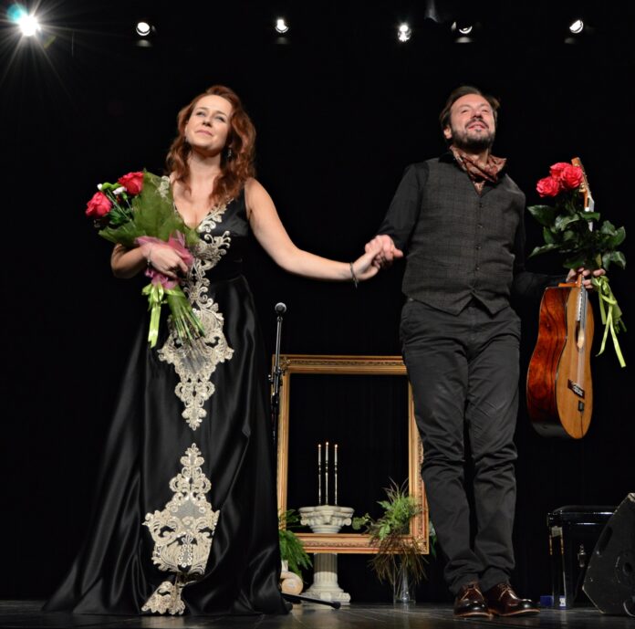 Na przyciemnionej scenie stoją kobieta (od lewej) i mężczyzna (po prawej). Kobieta ma czarną długą suknię z białym haftowanym wzorem na całej długości, w prawej ręce trzyma bukiet czerwonych róż. Ma długie rude kasztanowe, pofalowane włosy. Mężczyzna obok trzyma ją za rękę. Ubrany jest na czarno, ma zapinaną kamizelkę w kratę i brązową chustkę pod szyją. W prawej ręce trzyma gitarę i bukiet czerwonych róż. Uśmiecha się do publiczności