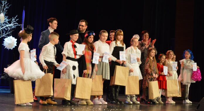 Na zdjęciu stoi grupa dzieci wraz z dorosłymi, uczestnikami konkursu na pomysłową p[piosenkę zimową. Dzieci ubrane się w większości na biało i trzymają torby z nagrodami w rękach. Za nimi stoją jurorzy konkursu oraz konferansjer