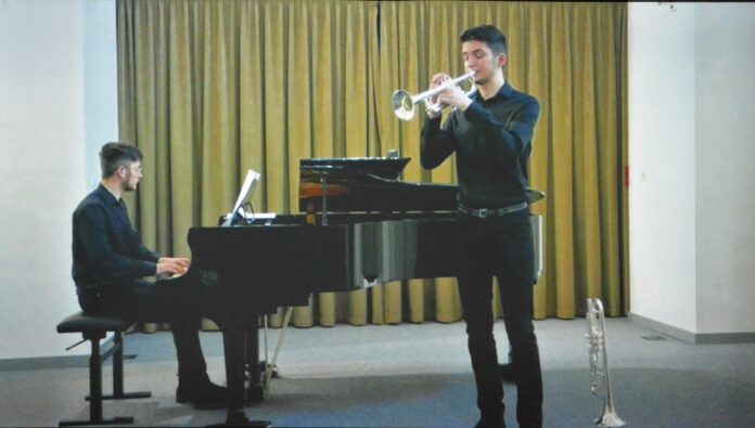 Obraz zawiera: dówch młodych mężczyzn na scenie. Jeden gra na fortepianie, drugi na trąbce.