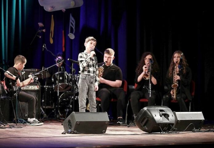 Obraz zawiera: Zespół Jazz Kids na scenie Cenrtum Kultury w Przemyślu. Na pierwszym planie chłopiec śpiewający do mikrofomu. Za nim siedzą muzycy saksofonista, trębaczka, saksofonistka. Z boku basista, a z tyłu perkusista.