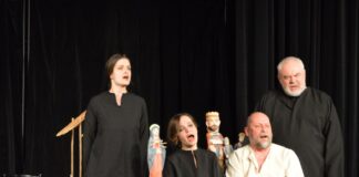 Na scenie cztery postacie: stojąca kobieta, obk siedzi kobieta, obok niej mężczyzna. Ostatni mężczyzna stoi obok siedzącego