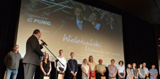 Historia pioniera gazownictwa na dużym ekranie Na scenie stoi grupa ludzi, statystów, biorących udział w filmie. Przed mikrofonem producent filmu, ubrany w szary garnitur w duża biała kratę. W tle obraz z filmu z napisami