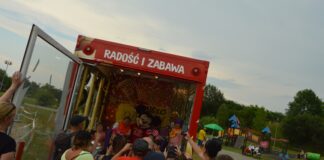 Wakacyjne Teatry Profilaktyczne: Disco bus z Myszką Miki Przed tylnym wejsciem do dużego czerwonego samochodu stoją dorośli ludzie, którzy chcą zrobić zdjęcia siedzącym w nim dzieciom. w tle widać pochmurne niebo, plac zabawa oraz zieleń