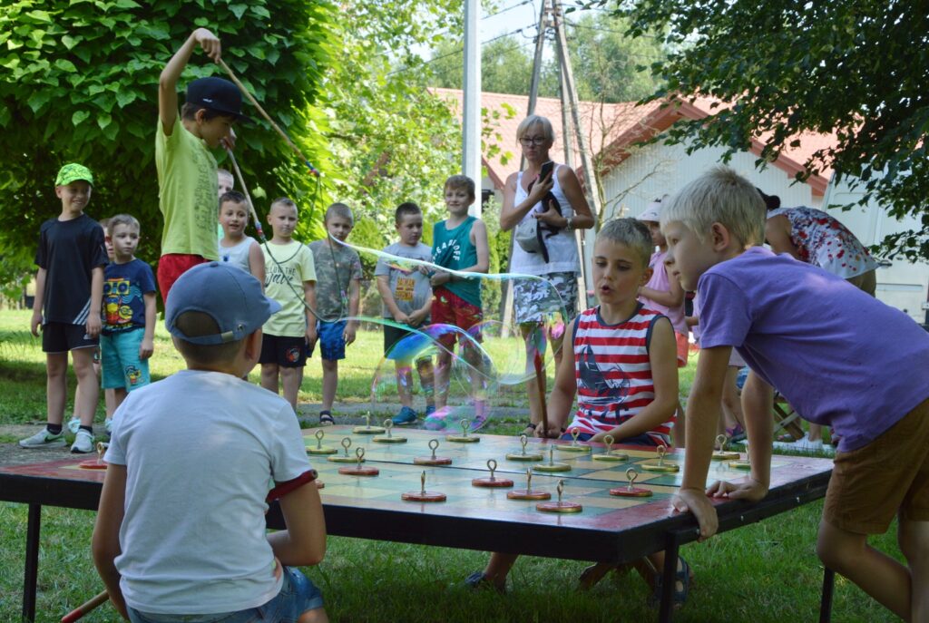 Przy dużym szachowym stole znajduje się trzech małych chłopców, nieopodal nich innych chłopiec robi duże banki mydlane. Przygląda się temu grupka dzieci oraz jedna kobieta