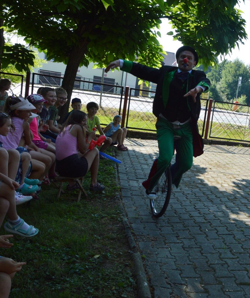 Klaun jedzie po chodniku na jednokołowym rowerze, bez kierownicy. Niedaleko siedzą dzieci i obserwują klauna