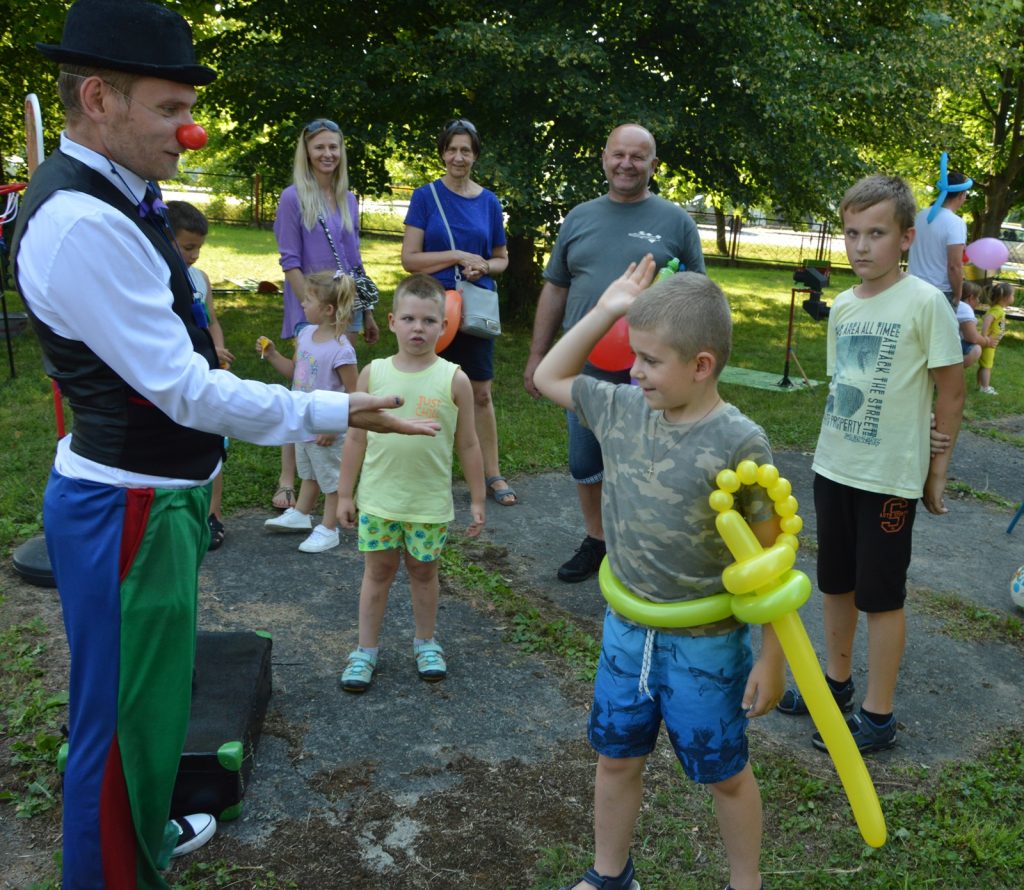 mały chłopiec przybija "piątkę" klaunowi w podziękowaniu za balon w kształcie szpady. Wokół stoją inne dzieci oraz dorośli.