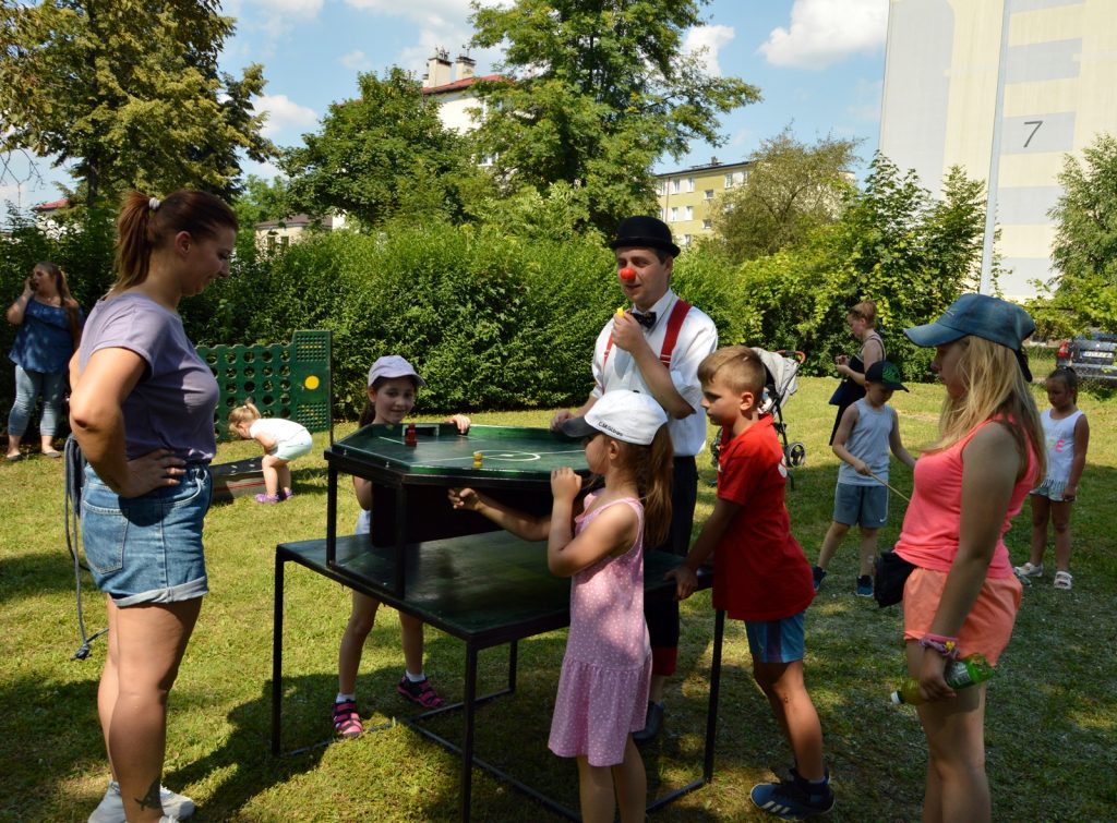 Przy dwupiętrowym stoliku do gry w piłkarzyki stoi troje dzieci oraz kobieta i klaun. Rozmawiają ze sobą i przygotowują się do gry