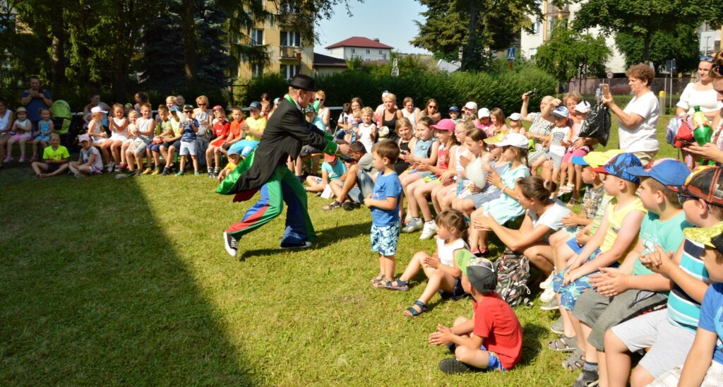Klaun podbiega do grupy dzieci i zaprasza do zabawy. Dzieci siedzą na ławkach