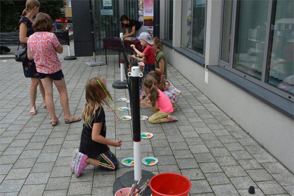 Dzieci maluija na czarnej folii kolorowymi farbami. W tle stanowisko Centrum Informacji Turystycznej i Kulturalnej.