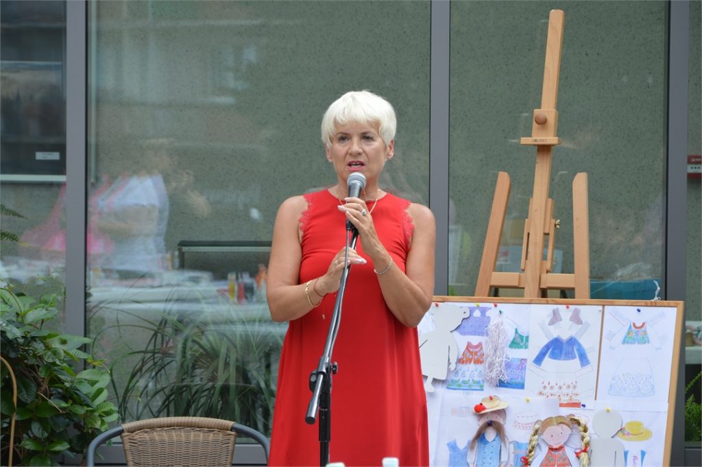 Dyrektor Jasielskiego Domu Kultury Helena Gołębiowska wita przybyłych na Weekend z Kulturą. Kobieta w czerwonej sukience, za nią sztaluga z pracami plastycznymi.