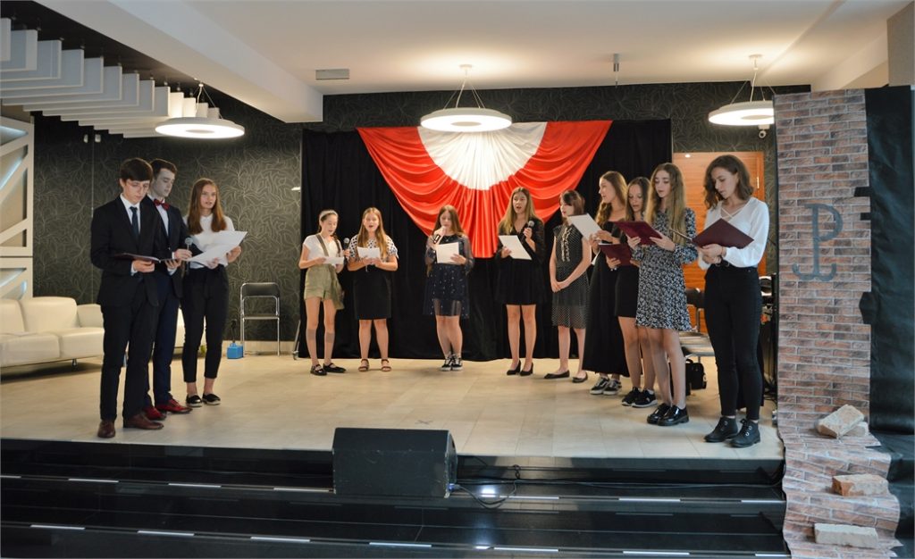 Młodzi recytatorzy i soliści JDK stoja w półokręgu na holu JDK i śpiewają powstańcze piosenki