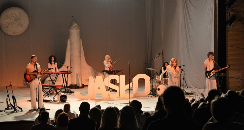 Muzycy i Ralph Kamiński na scenie. Na środku sceny wielki napis Jasło. W tle rakieta kosmiczna i księżyć.