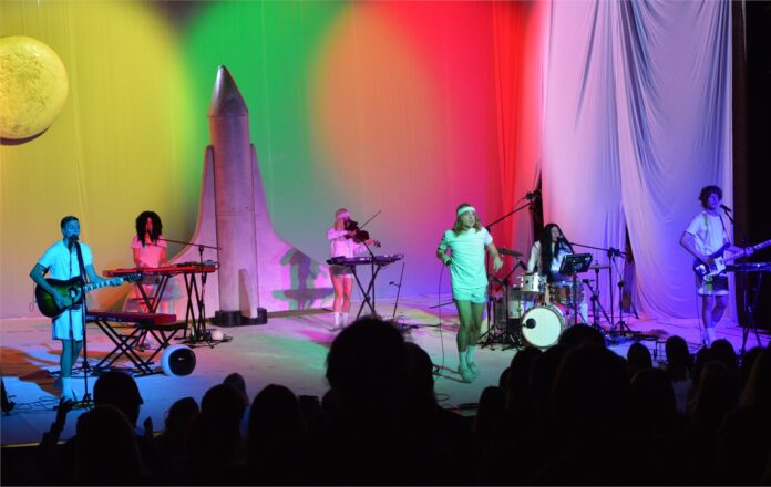 Ralph Kamiński na scenie JDK. Feeria barw. Z tyły rakieta kosmiczna i księżyc. Z instrumentami stoją Roger, Madeline, Samantha, Inez Przez Z oraz Laurent.