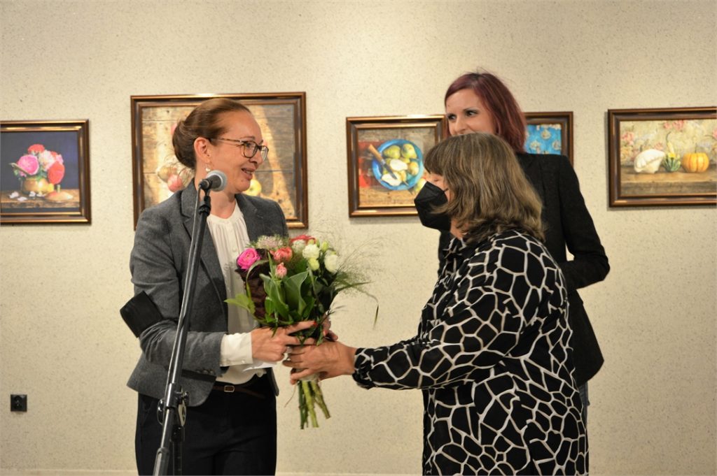 Ada Florek orrzymuje kwiaty od Anny Brożyny, plastyczki Jasielskiego Domu Kultury. Z tyłu Katarzyna Gądek Smyka. W tle obrazy artystki.