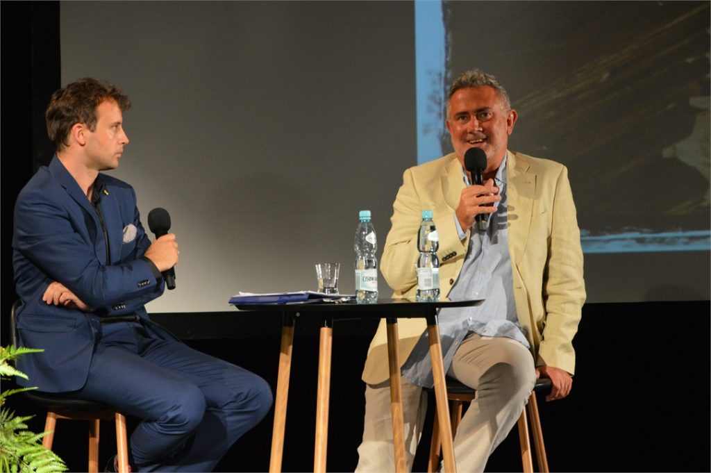 Andrezj Kurdziel rozmawia z Marcinem Kydryńskim. Dwóch mężczyzn siedzi na scenie JDK ptzy stoliku na ktorym jest szklanka i dwie butelki wody.