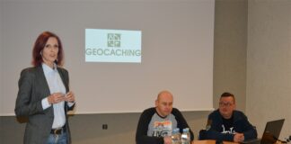 Pasja Geocachingu Obraz zawiera trzy osoby. Kobieta pierwsza od lewej stoi, z prawej strony za biurkiem siedzą dwa mężczyźni. Z tyłu umieszczony jest ekran projekcyjny