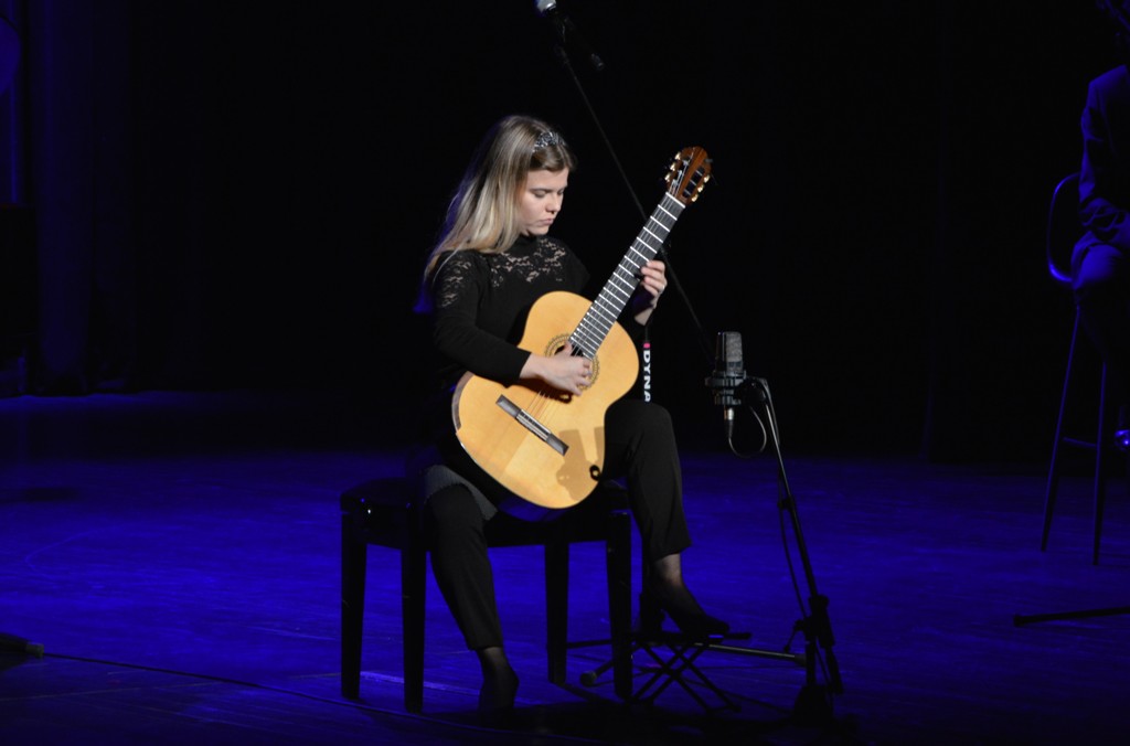Młoda kobieta siedzi na stołku na przyciemnionej scenie. Gra na gitarze. Obok niej stoi mikrofon.