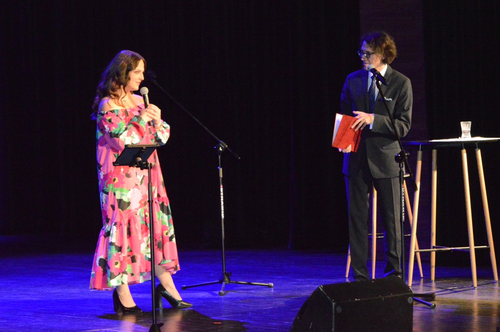 Z lewej strony sceny z mikrofonem w ręce Aleksandra Tocka, z praqwej strony sceny przed mikrofonem Adam Głaczyński. Rozmawiają ze sobą.