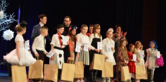 Koncert finałowy „Pomysłowej piosenki zimowej” Na zdjęciu stoi grupa dzieci wraz z dorosłymi, uczestnikami konkursu na pomysłową p[piosenkę zimową. Dzieci ubrane się w większości na biało i trzymają torby z nagrodami w rękach. Za nimi stoją jurorzy konkursu oraz konferansjer
