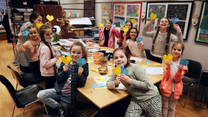Na zdjęciu grupa dziewczynek podczas zajęć plastycznych. Wszystkie pozują przy stole do zdjęcia, są zadowolone i uśmiechnięte. W dłoniach trzymają żółto niebieskie serduszka. Za nimi w tle na ścianie kolorowe prace plastyczne wykonane różnymi technikami