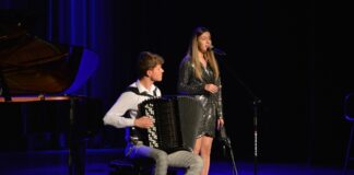 Duet z JDK doceniony w Międzynarodowym Festiwalu  Your Song Młody mężczyzna z akordeonem siedzi na stołku i akompaniuje młodej kobiecie śpiewającej do mikrofonu.