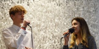 Duet z JDK na podium I Międzynarodowego Festiwalu Piosenki „My Song” Na białym pofalowanym tle, stoi dwie młode osoby. Po lewej stronie znajduje się młody mężczyzna, ubrany w białą koszulę i granatowe spodnie z brązowym paskiem. W prawej dłoni trzyma mikrofon, do którego śpiewa. Na głowie ma dużo kręconych, krótkich włosów. Naprzeciw niego na scenie stoi młoda dziewczyna . Ma dlugie4j jasnobrązowe włosy. Na szyi delikatny naszyjnik. Ubrana jest w ciemnoniebieską sukienkę z dekoltem, w drobne, granatowe kwiatuszki. W prawej dłoni trzyma mikrofon, do którego śpiewa, lewą trzyma w pasie. Ma na niej duży zegarek w kolorze srebra z ciemną tarczą