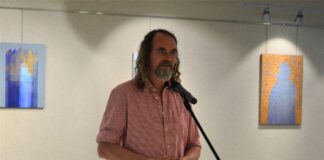Wyborski w JDK Obraz zawiera: Paweł Wyborski stoi przed mikrofonem na statywie i przemawia do przybyłych na tle swoich obrazów.