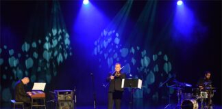 Bach w jazzowej odsłonie Na niebiesko oświetlonej scenie znajduje się trzech muzyków., ubranych na czarno. Jeden siedzi i gra na instrumentach klawiszowych, drugi stoi i gra na puzonie, trzeci siedzi i gra na perkusji.