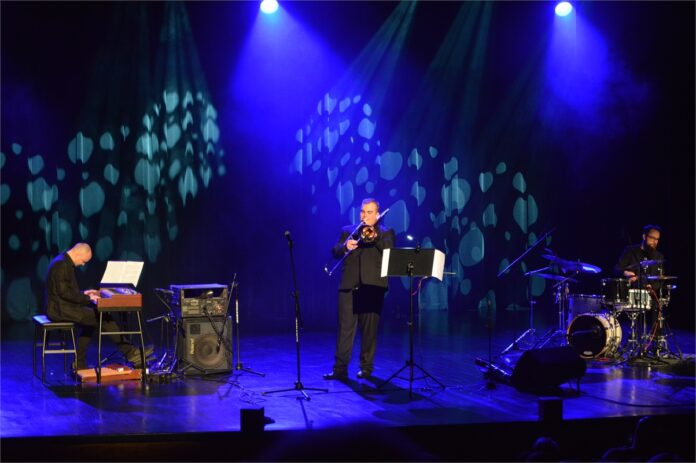 Na niebiesko oświetlonej scenie znajduje się trzech muzyków., ubranych na czarno. Jeden siedzi i gra na instrumentach klawiszowych, drugi stoi i gra na puzonie, trzeci siedzi i gra na perkusji.