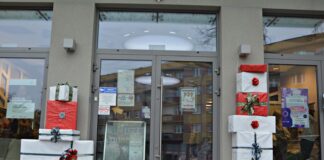 „Na prezenty czas” – kolorowo i świątecznie Na zdjęciu znajduje się przeszklone wejście do budynku,. Po obydwu stronach drzwi stoją zapakowane w prezenty duże pudła, przewiązane kokardą. Na drzwiach powieszone są male plakaty z zapowiedzią wydarzeń