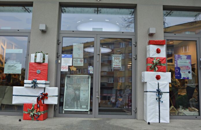 Na prezenty czas (17) Na zdjęciu znajduje się przeszklone wejście do budynku,. Po obydwu stronach drzwi stoją zapakowane w prezenty duże pudła, przewiązane kokardą. Na drzwiach powieszone są male plakaty z zapowiedzią wydarzeń