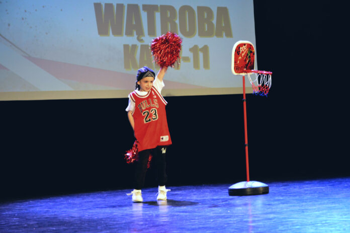 Obraz zawiera: Na scenie JDK dziewczynka w sportowym stroju z pomponami tańczy taniec nowoczesny. Z prawej strony obok niej stoi tablica z koszem.