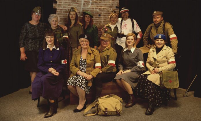 Dziewczyny z Powstania Obraz zawiera: Grupę osób w powstańczych ubiorach z opaskami na rękach. Z przodu siedzi pięć sanitariuszek. Z tyłu z lewej strony stoi instruktor muzyki JDK Ewa Grzebień w żołnierskiej czapce oraz szóstka powstańców.