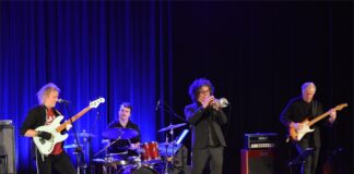Koncert muzycznych pejzaży Obraz zawiera: czterech muzyków na scenie JDK. Przed nim dwa głośniki odsłuchowe. Muzyk od lewej z długimi siwiejącymi włosami gra w specjalnych rękawiczkach na gitarze elektrycznej. Pomiędzy nim, a trębaczem, nieco z tyłu gra perkusista. Z prawej strony czwarty muzyk składu gra na gitarze basowej, która podpięta jest do stojącego za nim wzmacniacza.