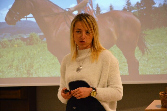 29. Klub Ludzi z Pasją. Konie_4 Obraz zawiera: na pierwszym planie gość klubu z pasją opowiada o swoich czworonożnych podopiecznych. Z tyły w tle ekran projektora na którym wyświetlone jest zdjęcie konia z jeźdźcem.