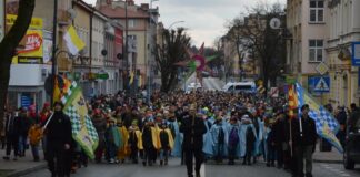 Obraz zawiera: Tłum idący ulicą Jasła. Na pierwszym planie niosący gwiazdę, po jego lewej i prawej stronie harcerze z orszakowymi chorągwiami. Za nim Trzej Królowie oraz uczniowie z jasielskich szkół w pelerynach i z koronami na głowie. Za nimi pozostali uczestnicy orszaku.