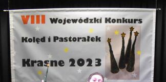 Sukcesy kolędowe wokalistów JDK Obraz zawiera: Młody chłopak w trakcie podskoku, z dyplomem w lewej ręce i statuetką w prawej. Za nim z tyłu sceny wisi baner na którym znalazło się zdjęcie czterech statuetek z napisem: Ósmy Wojewódzki Konkurs Kolęd i Pastorałek - Krasne 2023.