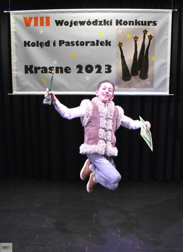 FB_IMG_1673537276401 Obraz zawiera: Młody chłopak w trakcie podskoku, z dyplomem w lewej ręce i statuetką w prawej. Za nim z tyłu sceny wisi baner na którym znalazło się zdjęcie czterech statuetek z napisem: Ósmy Wojewódzki Konkurs Kolęd i Pastorałek - Krasne 2023.