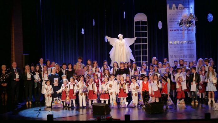 P1020477 Obraz zawiera: Wszyscy finaliści Koncertu "Na Nutę Kantyczek - Kolędy Podkarpacia", stoją na scenie wraz z jurorami, mecenasami kultury, dyrektor JDK oraz zastępcą burmistrza Jasła, Elwirą Musiałowicz - Chech i śpiewają wspólnie kolędę. Za nimi scenografia koncertu czyli wielki, biały anioł, duże podłużne okno, duże płatki śniegu oraz z prawej strony zwisający z góry baner z napisami "Na nutę kantyczek".