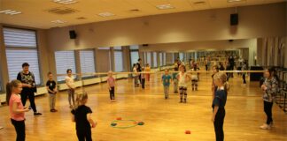 Obraz zawiera: dzieci wraz z instruktorką stoją dookoła kółka do którego należy trafić woreczkami wypełnionymi groszkiem. W tle lustro, którym wyłożona jest cała ściana sali tanecznej.