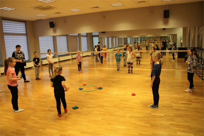 Zajęcia Wokalno - Ruchowe w JDK _4 Obraz zawiera: dzieci wraz z instruktorką stoją dookoła kółka do którego należy trafić woreczkami wypełnionymi groszkiem. W tle lustro, którym wyłożona jest cała ściana sali tanecznej.