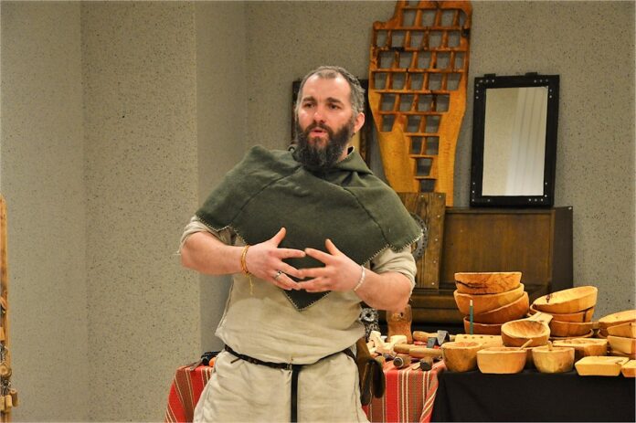 Obraz zawiera: Mężczyzna w średnim wieku w średniowiecznym stroju stoi na tle własnoręcznie wykonanych talerzy, misek i narzędzi z drewna. Na dalszym planie drewniana półka na przyprawy i lustro w drewnianej oprawie