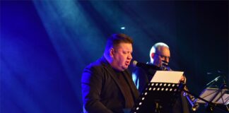 Obraz zawiera: piosenkarz Marek Dyjak siedzi na krześle, przed nim pulpit z tekstami piosenek i śpiewa do mikrofonu. Obok, stojąc gra saksofonista.