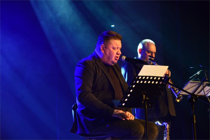 Marek Dyjak_3 Obraz zawiera: piosenkarz Marek Dyjak siedzi na krześle, przed nim pulpit z tekstami piosenek i śpiewa do mikrofonu. Obok, stojąc gra saksofonista.