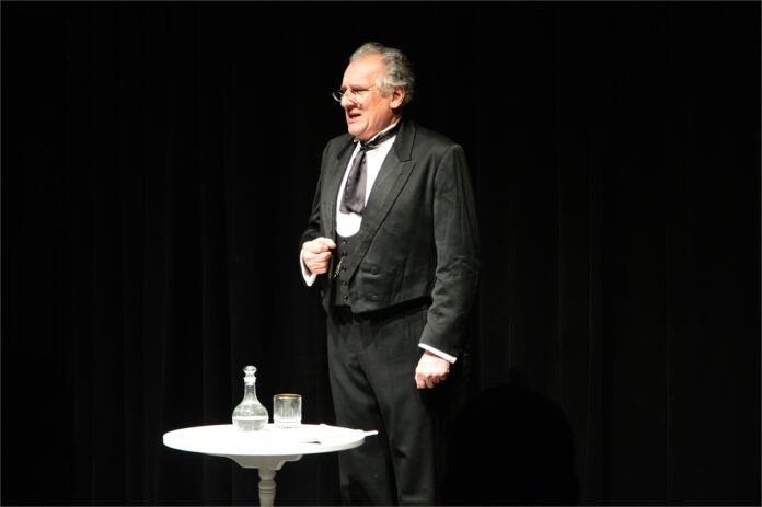 SK JDK BWA Sanok_3 Obraz zawiera: Siwiejący mężczyzna w okularach, a także w surducie, spodniach i kamizelce z XIX wieku stoi przodem do widowni. To aktor z monodramu Czechowa. Przed nim stoi stolik z karafką wody, a obok szklanka.