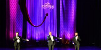 Obraz zwiera: Trzech mężczyzn stoi na scenie Jasielskiego Domu Kultury. Wszyscy mają w rękach mikrofony i śpiewają. Nad nimi wisi przystrojony szarfą żyrandol z kilkunastoma żarówkami w kształcie świeczek. Z tyłu zwisają tiulowe elementy dekoracji.