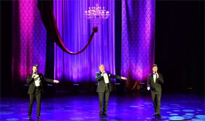 Pieśni z miłością w tle Obraz zwiera: Trzech mężczyzn stoi na scenie Jasielskiego Domu Kultury. Wszyscy mają w rękach mikrofony i śpiewają. Nad nimi wisi przystrojony szarfą żyrandol z kilkunastoma żarówkami w kształcie świeczek. Z tyłu zwisają tiulowe elementy dekoracji.