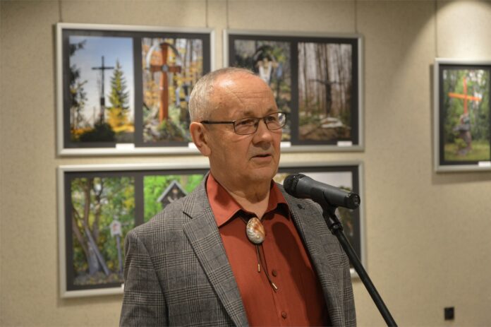 31. Klub Ludzi z Pasja_3 Obraz zawiera: Na pierwszym planie siwy mężczyzna około 60 -ciu lat w okularach w marynarce w jodełkę stoi przed mikrofonem i patrzy przed siebie. Z tyłu na ścianach w dużych ramach fotosy przydrożnych krzyży jego autorstwa.