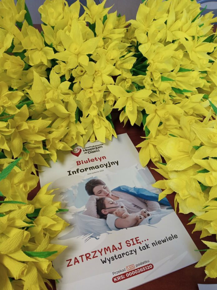 -5669279351269892025 Sztuczne żonkile leżą na stole a po środku nich znajduje się biuletyn informacyjny dotyczący działalności podkarpackiego hospicjum dla dzieci. Biuletyn ma białą okładkę, a na niej czerwone i czarne napisy. Po środku zdjęcie kobiety z chłopcem leżącym na łóżku.