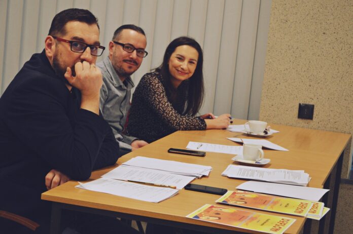 Na zdjęciu jest komisja konkursowa. Od lewej siedzi mężczyzna ubrany ciemną marynarkę, okulary w ciemnej oprawie, siedzi podparty i patrzy w obiektyw aparatu. Obok niego drugi mężczyzna w niebieskiej koszuli, ma okulary w ciemnej oprawie i ciemne włosy. patrzy w obiektyw aparatu i uśmiecha się. Jako ostatnia, przy ścianie siedzi młoda kobieta o ciemnych włosach i promiennym uśmiechu. Patrzy w obiektyw aparatu